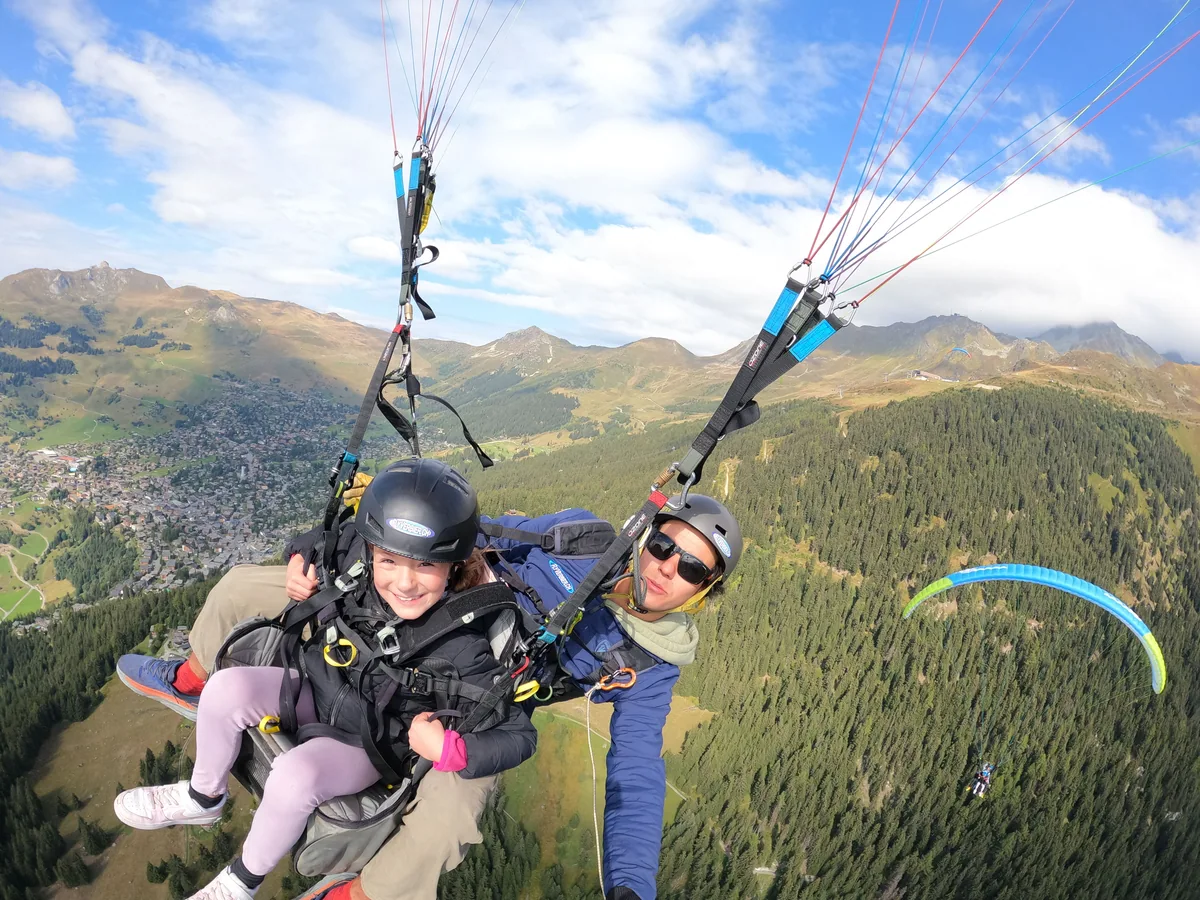 Une passagère souriante en parapente biplace au-dessus du village et des forêts